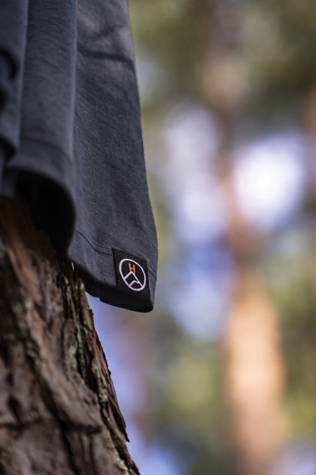 A close-up of a dark charcoal gray long-sleeve merino shirt hanging from a tree trunk outdoors, featuring a small circular woven label logo patch on the hem