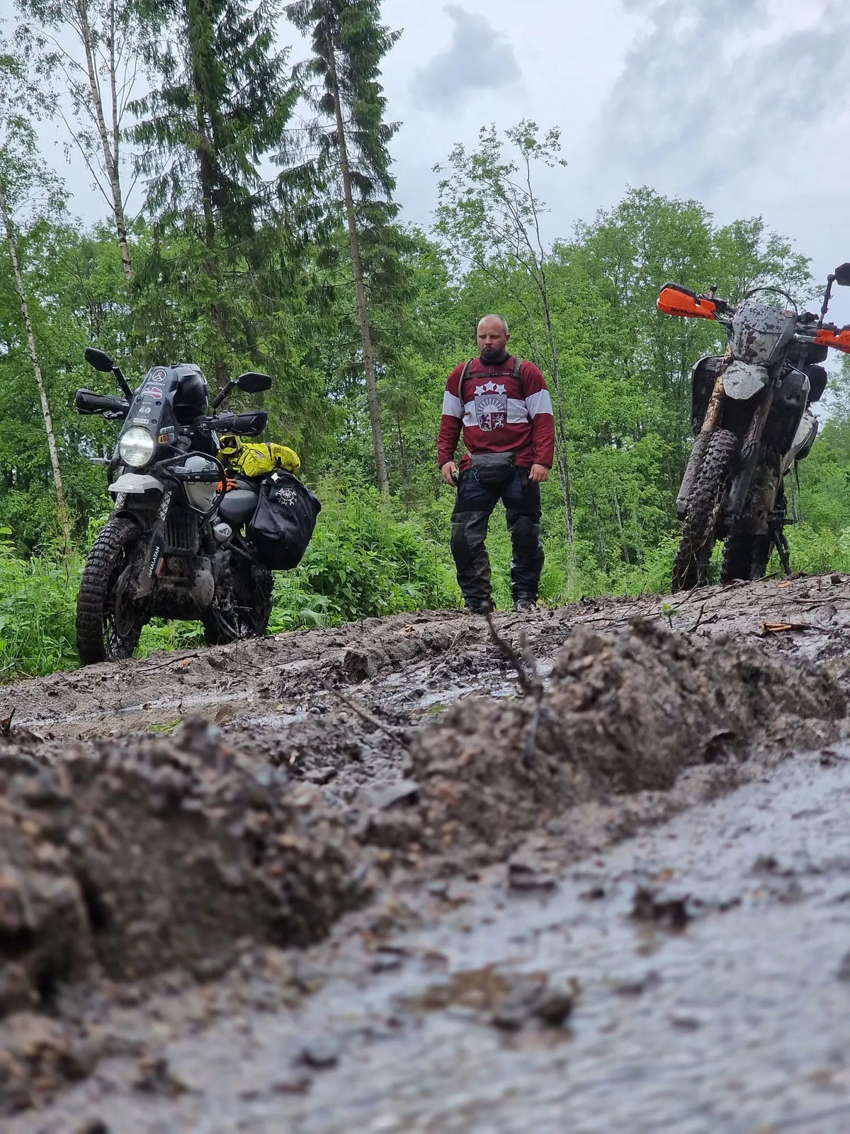 HEMCROFT Founder stands on a muddy forest road between two off-road motorcycles, with trees and an overcast sky in the background.