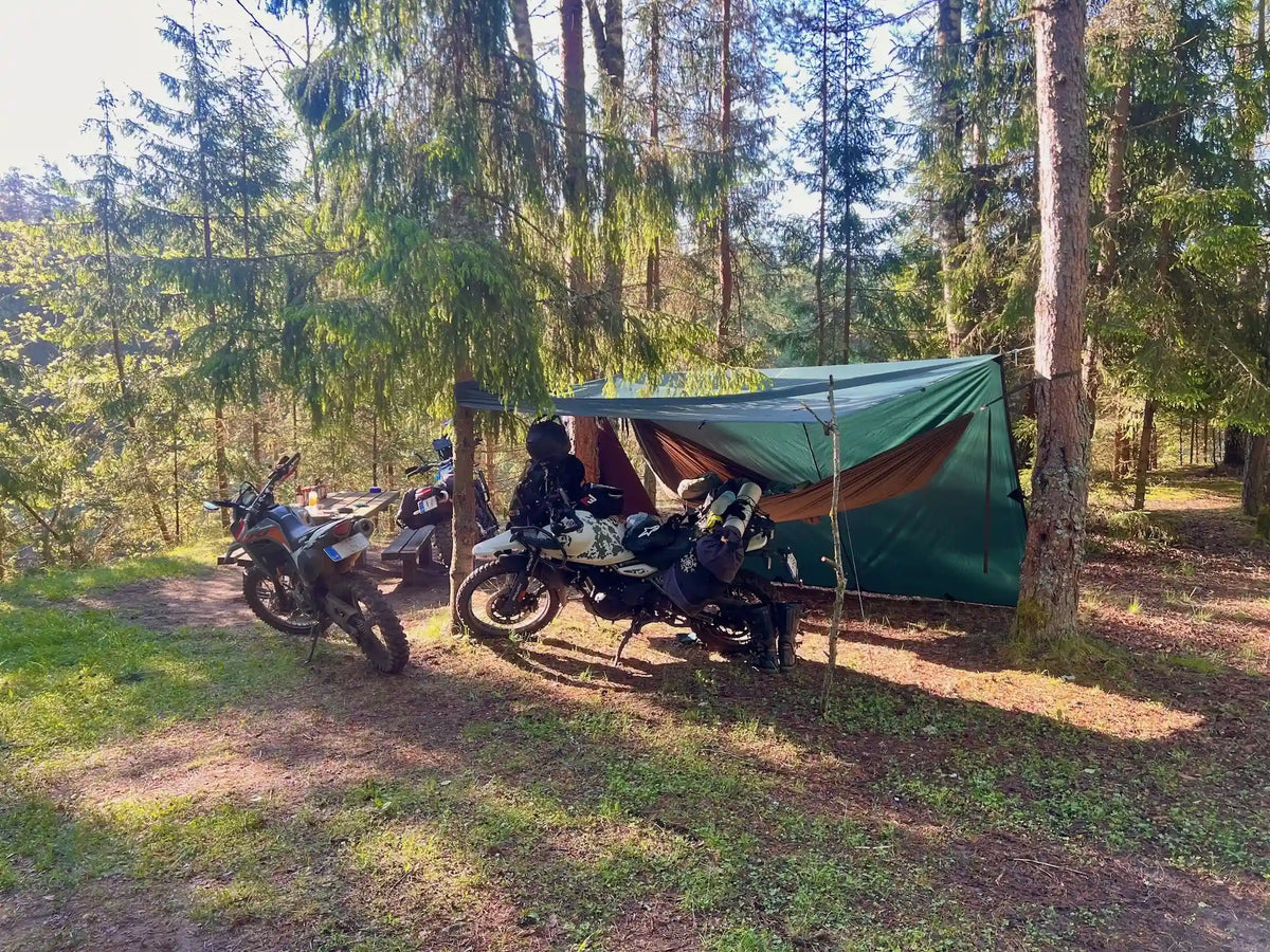 A green camping tarp shelter set up in a forest clearing with three motorcycles parked underneath and beside it, surrounded by tall pine trees.